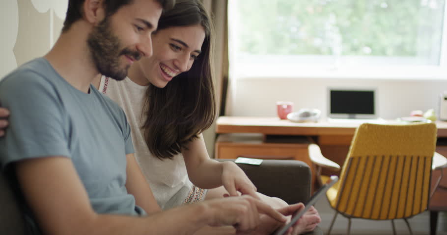 Young couple shopping online using credit card payment purchasing on digital touch screen together sitting on sofa celebrating with a kiss