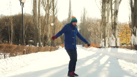 young man with beard in blue coat, jeans and colorful hat active dances in front of camera and glaze at camera while walking in the snowy winter park under the sunlight