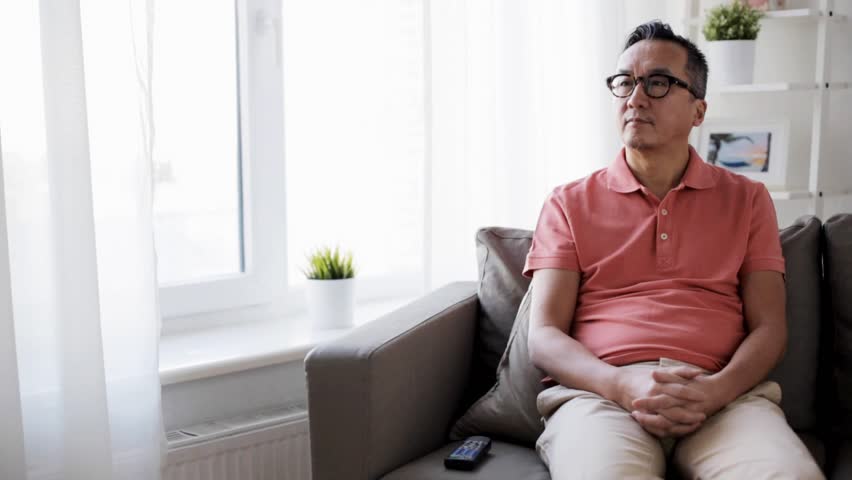 people, television and entertainment concept - man in eyeglasses with remote control watching tv at home