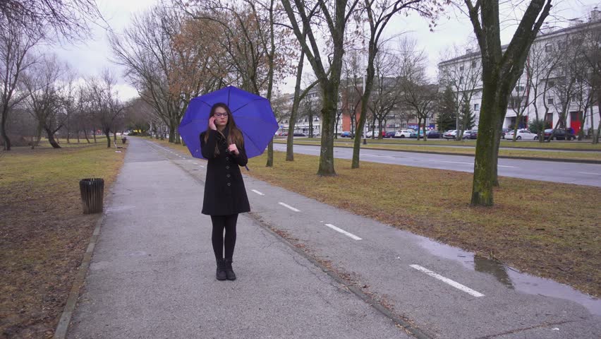 Girl with glasses and umbrella walking on rainy day and talking on the smartphone. She is upset and in negative mood. Passing by the camera, camera tracking her.