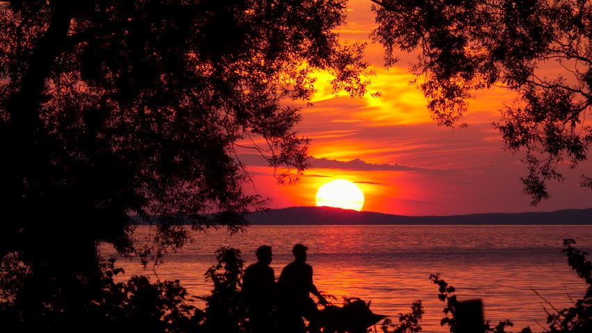 Silhouette of two people walking in front of orange sunset over lake. Gardening work, pushing wheelbarrow at dusk.