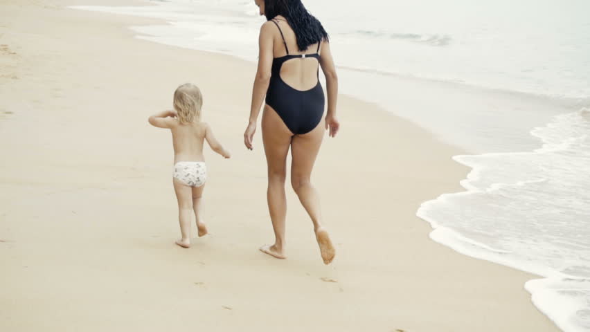 Rear view of young pretty woman with african braids in swimsuit and her little daughter are running on the beach and having fun