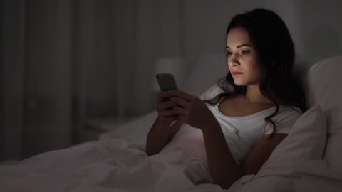 people, bedtime and rest concept - young woman in bed with smartphone going to sleep at home