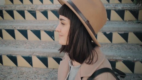 Camera moves all over girl in hat and coat,sitting on old rustic stairs ,listening favorite music on smartphone through white earbuds,smiling,looks around and slightly moves in tempo.Close up portrait
