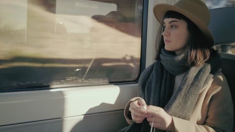 Short haired girl in hat travels in train, puts headphonesand in her ears, turns on music on iphone and smiles listening song, looking through window and browsing playlist, close up view