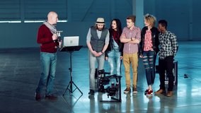 Group of filmmakers using handheld camera gyro stabilizing gimbal. Crew of young men and women watching the director showing camera man some moves standing in empty studio hall.