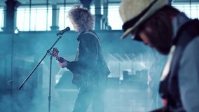 Rock band with curly young woman lead singer performing in empty hall, singing and dancing while shooting music video with smoke. Side view with male guitar player in foreground. Movement , round shot.