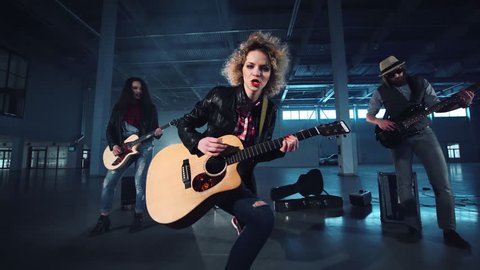 Curly haired woman in black jacket playing acoustic guitar in rock band, singing and dancing with her band members performing standing on sides, camera moving forward and back. Wide shot.
