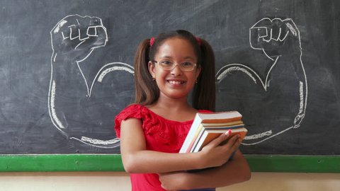 Concept on blackboard at school. Young people, student and pupil in classroom. Intelligent and successful hispanic girl in class. Portrait of female child smiling, looking at camera, holding books
