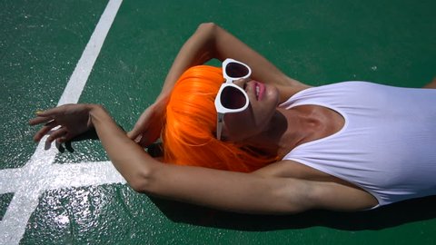 Closeup face of sexy woman in modern futuristic style lying on her back isolated on green play field. Creative look of woman wearing white swimsuit, sunglasses and orange wig in hot summer day