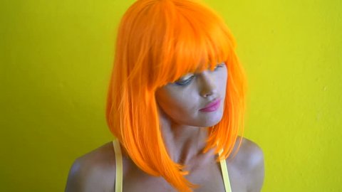 Closeup portrait of woman with colourful makeup in futuristic style shaking her hair and smiling over yellow wall background. Creative look of woman in yellow bra and orange wig - slow motion video