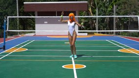 Happy woman running, jumping and looking into the camera with headphones on play field over tropical background. Creative look of woman in white swimsuit, sunglasses and orange wig - slow motion video