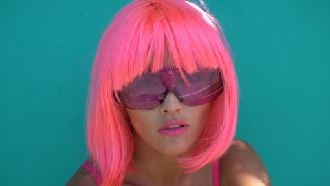 Closeup portrait of woman in futuristic style with purple sunglasses looking sensually into the camera over green mint wall background. Creative look of woman in pink bra and wig - slow motion video