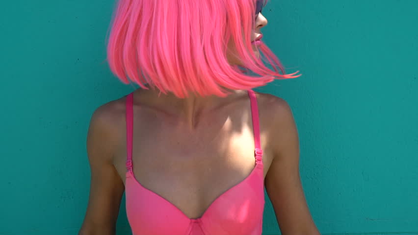 Closeup portrait of sexy woman with purple sunglasses looking into the camera and shaking her hair over green mint wall background. Creative look of woman in pink bra and wig - fast motion video