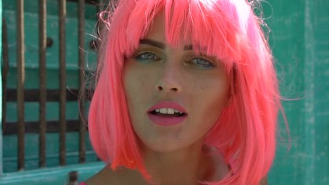 Closeup portrait of woman with colourful makeup wearing pink bra and wig looking into the camera, sending air kiss and smiling during summer day over green wooden door background - slow motion video