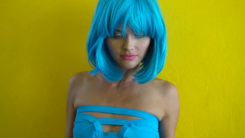 Closeup portrait of sexy woman with colourful makeup smiling, clapping hands and looking into the camera over yellow wall background. Creative look of woman in blue bikini and wig - slow motion video

