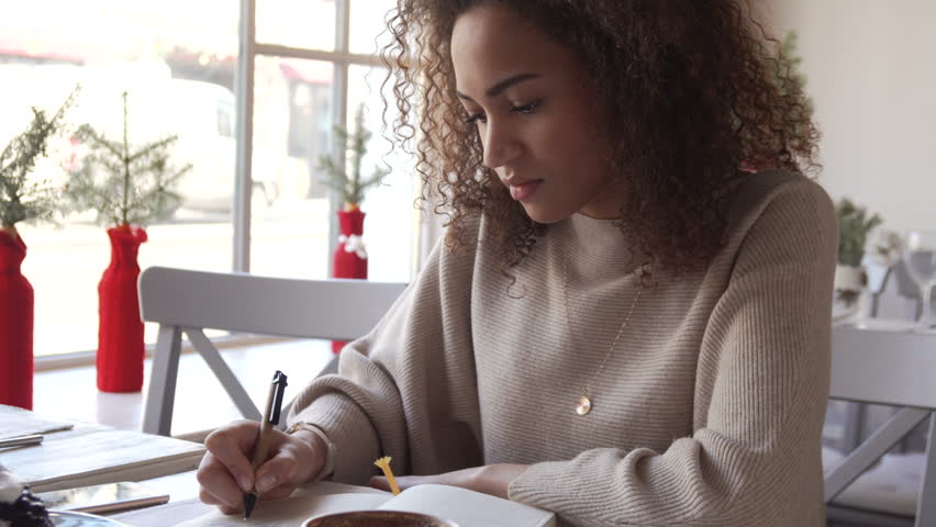 young afro amarican woman with curly hair make sketch and write notes in her notebook with coffee sitting alone in cafe near the window during sunny day