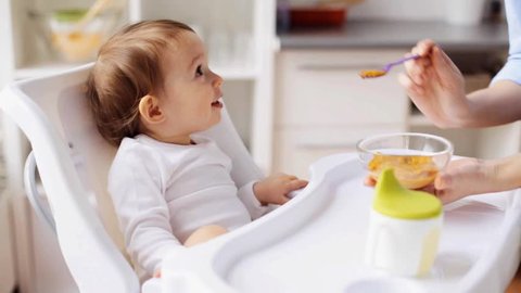 mother trying to feed little baby with fruit or vegetable mash at home kitchen