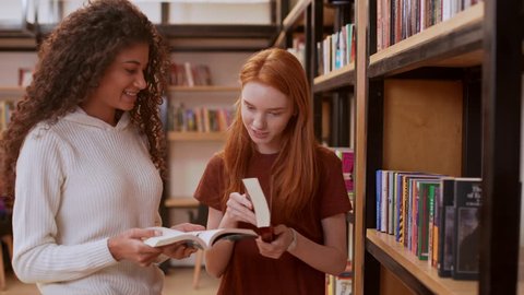 Young beautiful girl with curly hair and white sweater discussing books with female redhead teenage in library smiling
