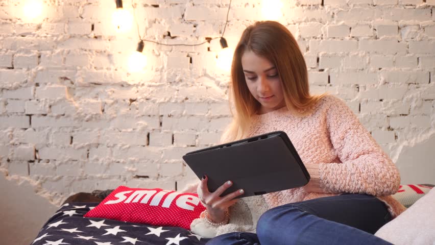 beautiful young girl chatting in internet on a sofa with soft pillows and garland on the wall.