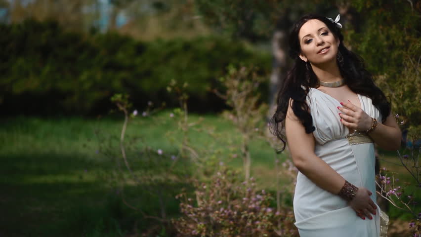 Young brunette caucasian woman in white dress pose near the blooming bush. Lady get pleasure being in the nice place under soft sunlight. Springtime concept.