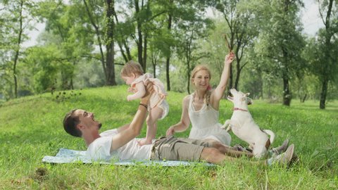 SLOW MOTION, CLOSE UP, DOF: Smiling young dad lying on blanket, playing having fun with cheerful sweet baby daughter. Beautiful mum sits beside and plays with cute little pet dog chasing a stick