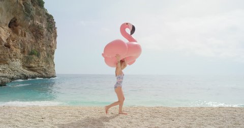 Woman walking on empty beach holding giant inflatable flamingo enjoying summer vacation on tropical beach holiday wearing full one piece swimsuit