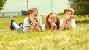 Three cute kids lying on the grass and looking playfully through magnifying glass