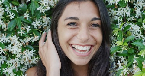 Close up portrait of beautiful young woman running hand through hair smiling in front of wall of flowers outdoors slow motion Adlı Stok Video