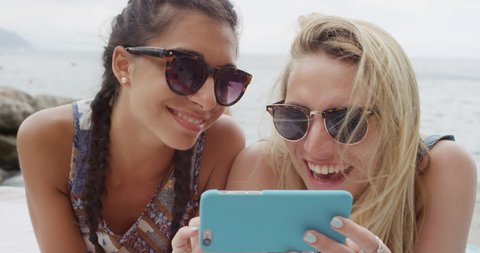 Two teenage girl friends hanging out at beach using mobile phone sending text message snapchat sharing digital content on social media enjoying summer vacation