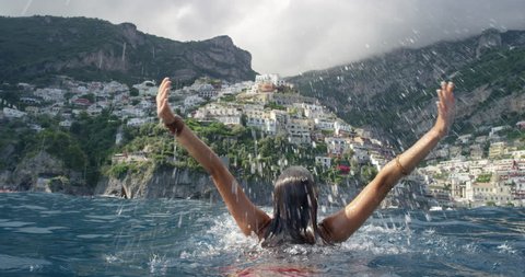Young tourist woman jumping out of water with arms up looking at Positano town in background Swimmer girl Celebrating Italian Vacation enjoying European summer holiday travel adventure in Amalfi Italy