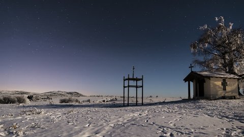 Night timelapse of small church with big tree and stars during full moon in winter season. Steps in the snow and bushes in foreground.
