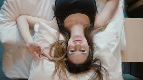 Happy young woman falls on the bed in room. Beautiful girl smiling, opens her eyes and looking to the camera.