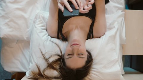 Young woman lying on the bed and holding smarphone. Girl browsing the Internet, chatting with someone.