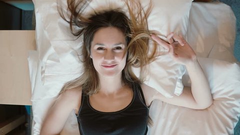 Happy young woman falls on the bed. Girl smiling, opens her eyes and looking to the camera.