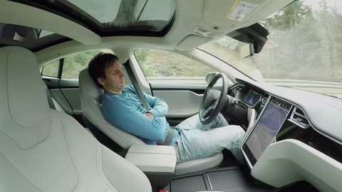 Male driver sleeping behind the self-driving steering wheel of an autonomous autopilot driverless car. Man fell deeply asleep while driving along the countryside road in luxury all-electric vehicle