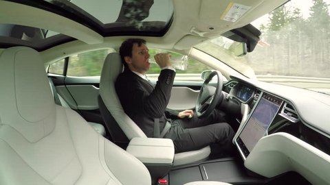 CLOSE UP: Irresponsible young businessman drinking alcohol behind the wheel while traveling in autonomous self-driving autopilot luxury electric driverless car. Male driver sipping vodka while driving