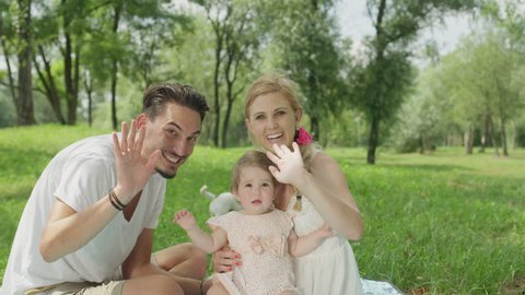 SLOW MOTION, CLOSE UP, DOF: Smiling Caucasian family enjoying quality time in park, waving into camera. Proud young father and pretty smiling mother sitting with cheerful child and having a video call