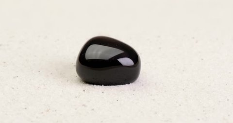 Obsidian gem rotating on white sand surface. Semi precious gemstone. Clip contains seamless loop.