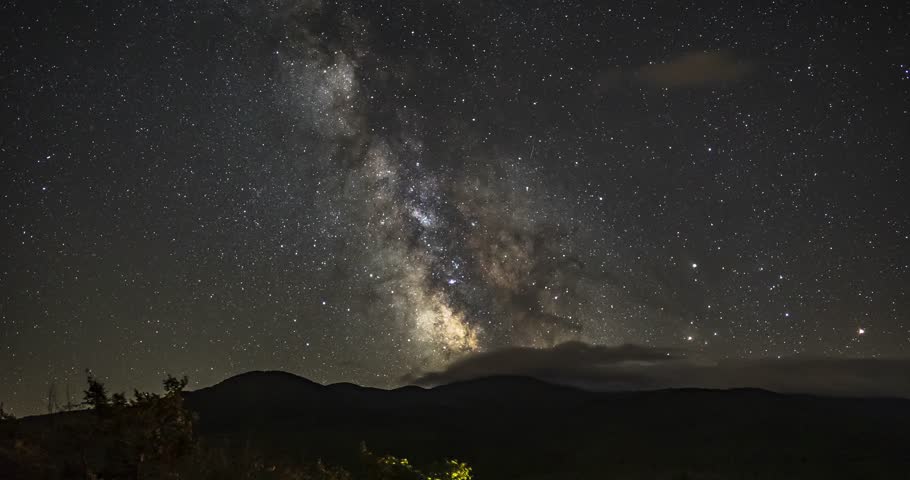 Night time-lapse of a starry sky and the milky way galaxy moving across the silhouette of mountains, while clouds circle around, in New Hampshire, USA.
