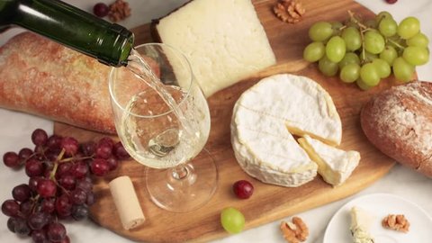 White wine poured into a glass at a tasting, with various types of cheeses, bread, and grapes