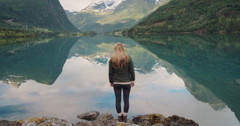 Woman standing at edge of lake looking at mountain view reflection in water Hiker girl wearing green down jacket scenic landscape enjoying vacation travel adventure nature Norway