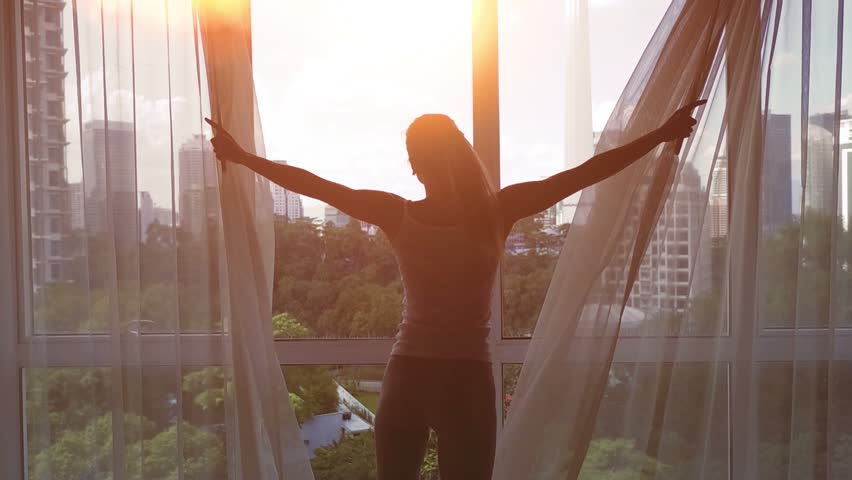 Slow motion. Young woman uncover the big window and looking out her apartment on the city buildings. Sunrise in the city.