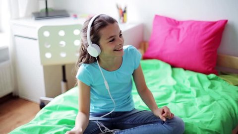 people, children, technology and leisure concept - happy girl with smartphone and headphones sitting on bed and listening to music at home