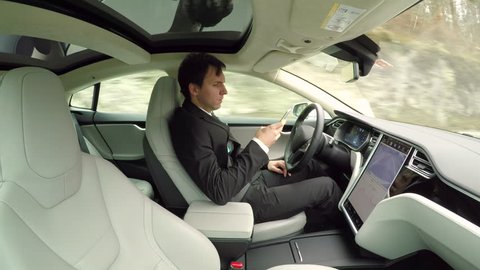 CLOSE UP: Young businessman texting writing messages on mobile phone while sitting behind self-driving steering wheel in autonomous autopilot driverless electric car traveling along countryside road