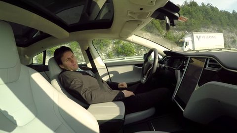 AUTONOMOUS TESLA CAR, FEBRUARY 2016:  Businessman sleeping behind the self-driving steering wheel of an autonomous electric Tesla car. Man fell deeply asleep while driving along the countryside road