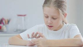 7 years old girl with smartphone. Girl sits at the table and uses smartphone, looking at camera and smiling.