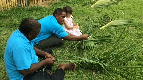 Indigenous Fijian men teach young tourist girl how to create a basket from weaving a Coconut Palm leaves.Travel Fiji concept, Real people. Copy space