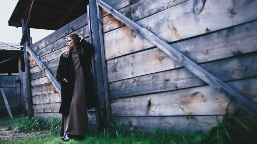 A young girl in a long beige dress posing against the backdrop of a village house