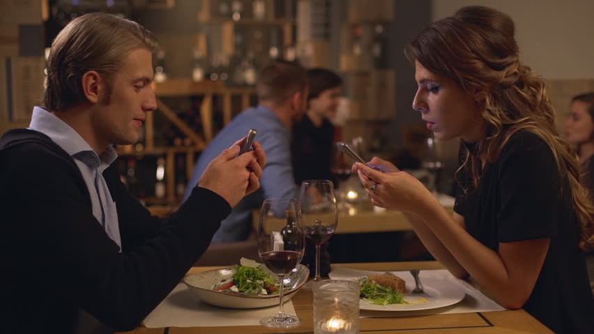 Two concentrated young Caucasian male and female sitting at restaurant opposite each other with mobile phones. Group of people are on background in slowmotion